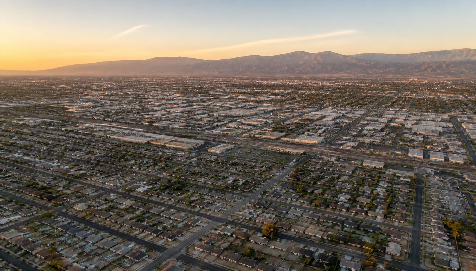 Aerial golden-hour view over a dense flat grid of modest homes and light-industrial blocks with a mountain range behind