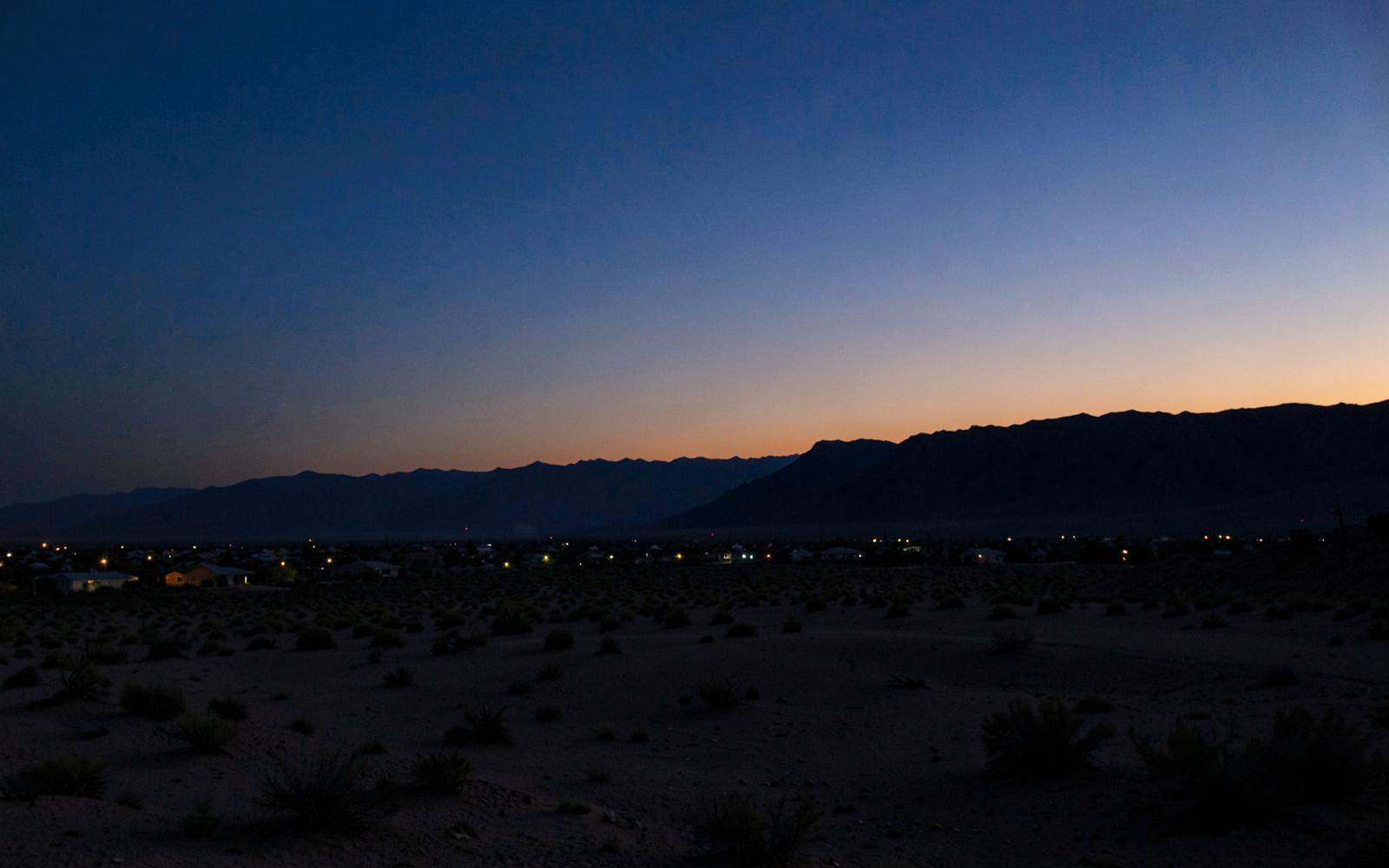 Wide desert basin at dusk with scattered distant house lights below a dark mountain silhouette and orange horizon