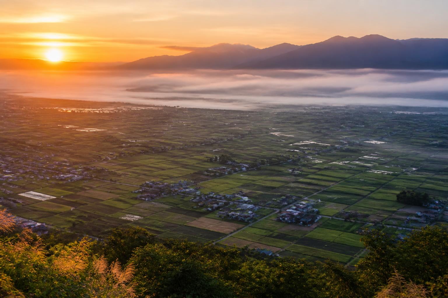 Elevated overlook of a wide agricultural valley at sunrise with patchwork fields, low mist and mountains beyond