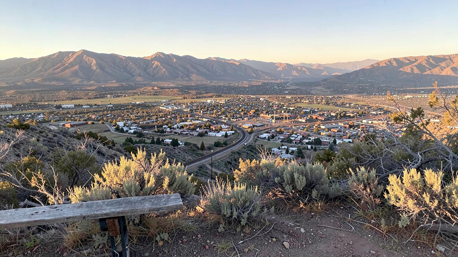 Overlook from a sagebrush hillside above a small city on a valley floor with rail lines crossing it and bare ranges behind