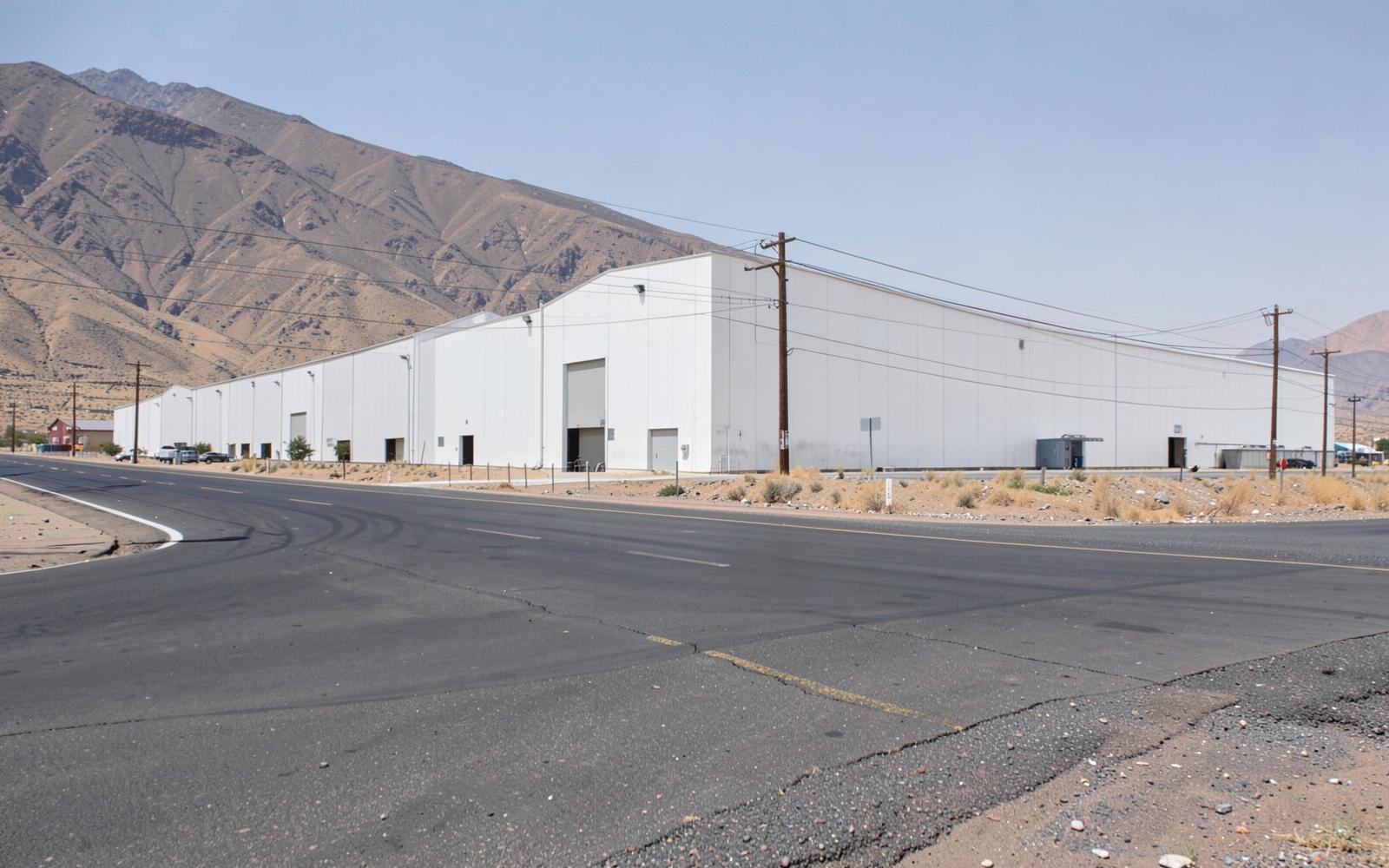 Row of large blank-walled distribution warehouses beside a wide industrial road in a desert valley