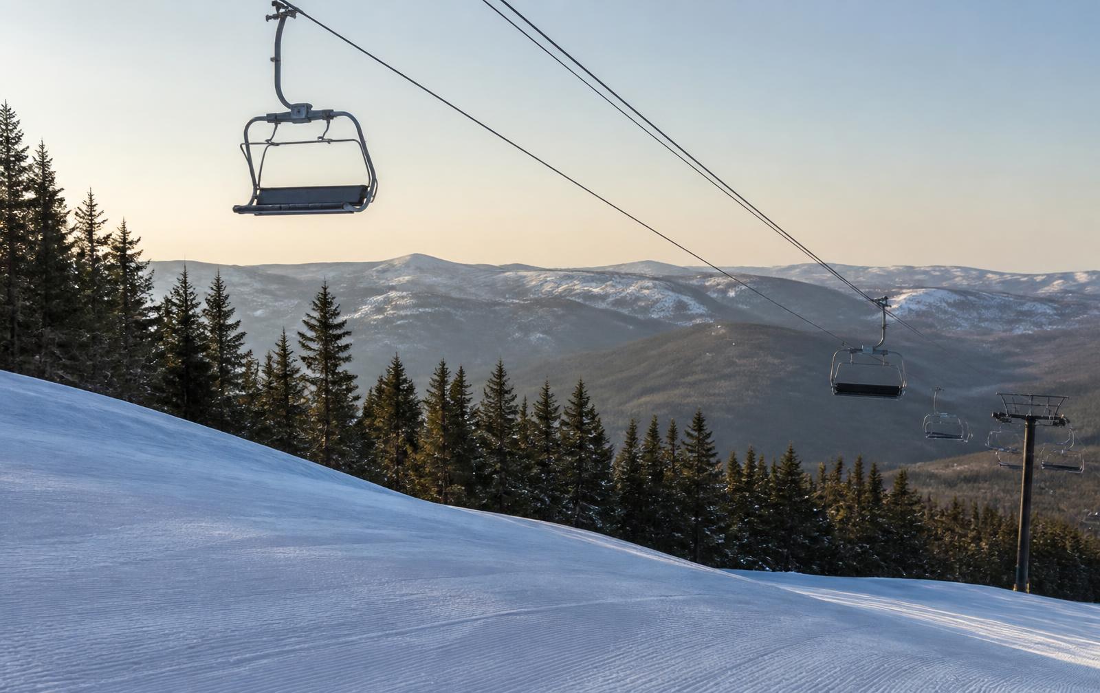 Empty ski lift chairs crossing above a groomed snowy slope with forested ridgelines in the distance