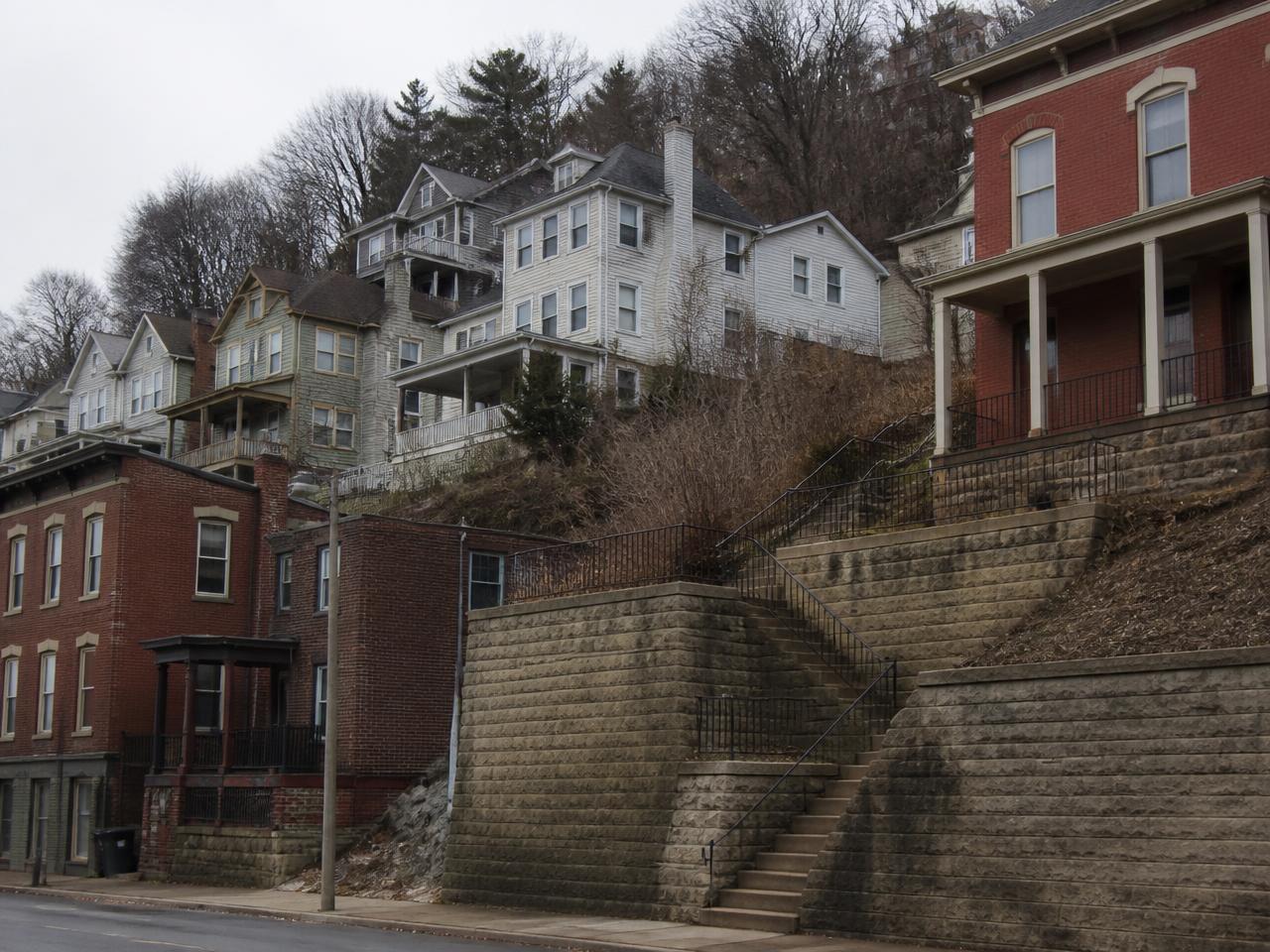 Older Pittsburgh houses stacked up a steep hillside above stone retaining walls and public steps