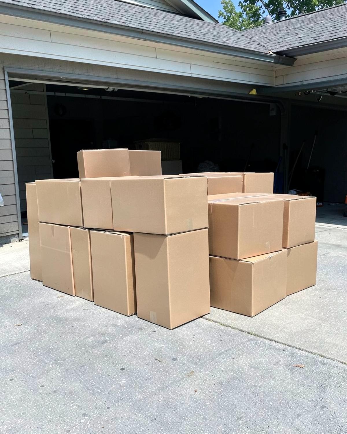 Plain cardboard moving boxes stacked on a suburban driveway in front of an open garage on a sunny day