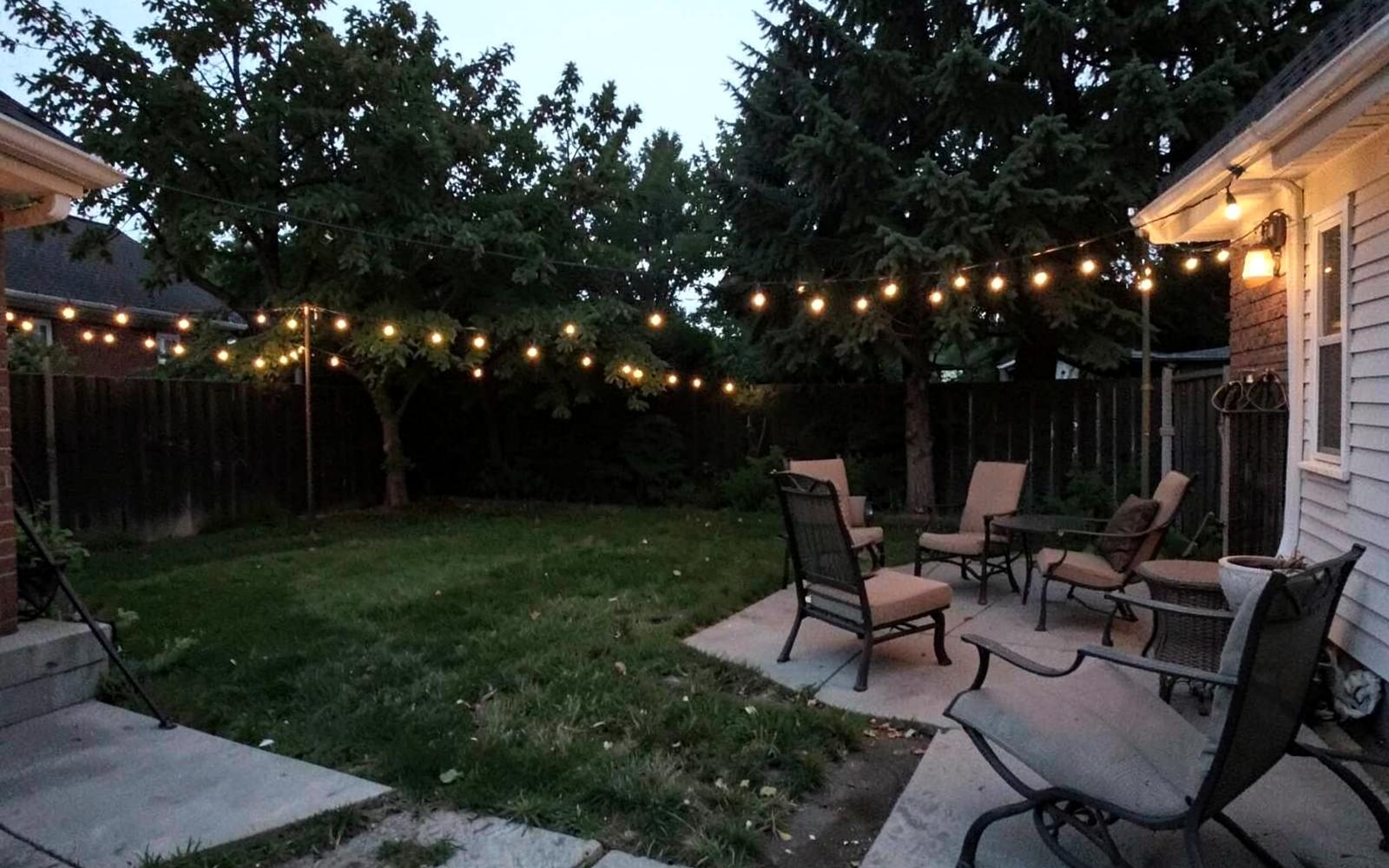 Suburban backyard patio at dusk with string lights glowing above chairs and a mown lawn