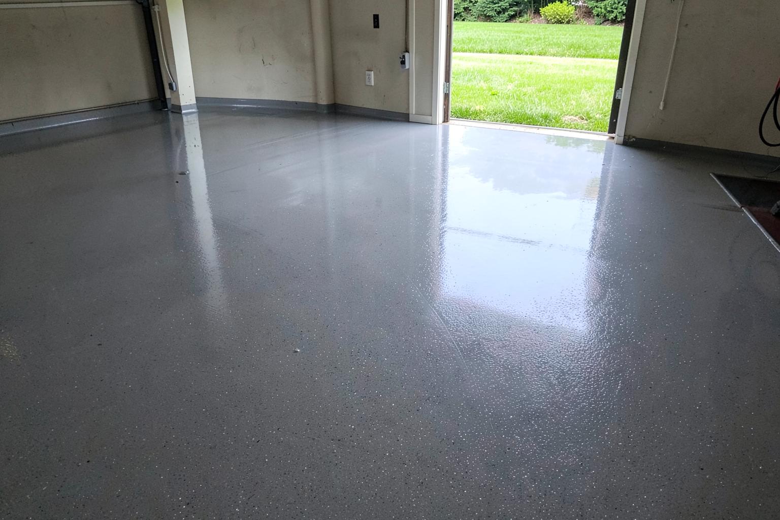 Freshly coated glossy grey flake epoxy garage floor in an empty garage with the side door open to a green lawn