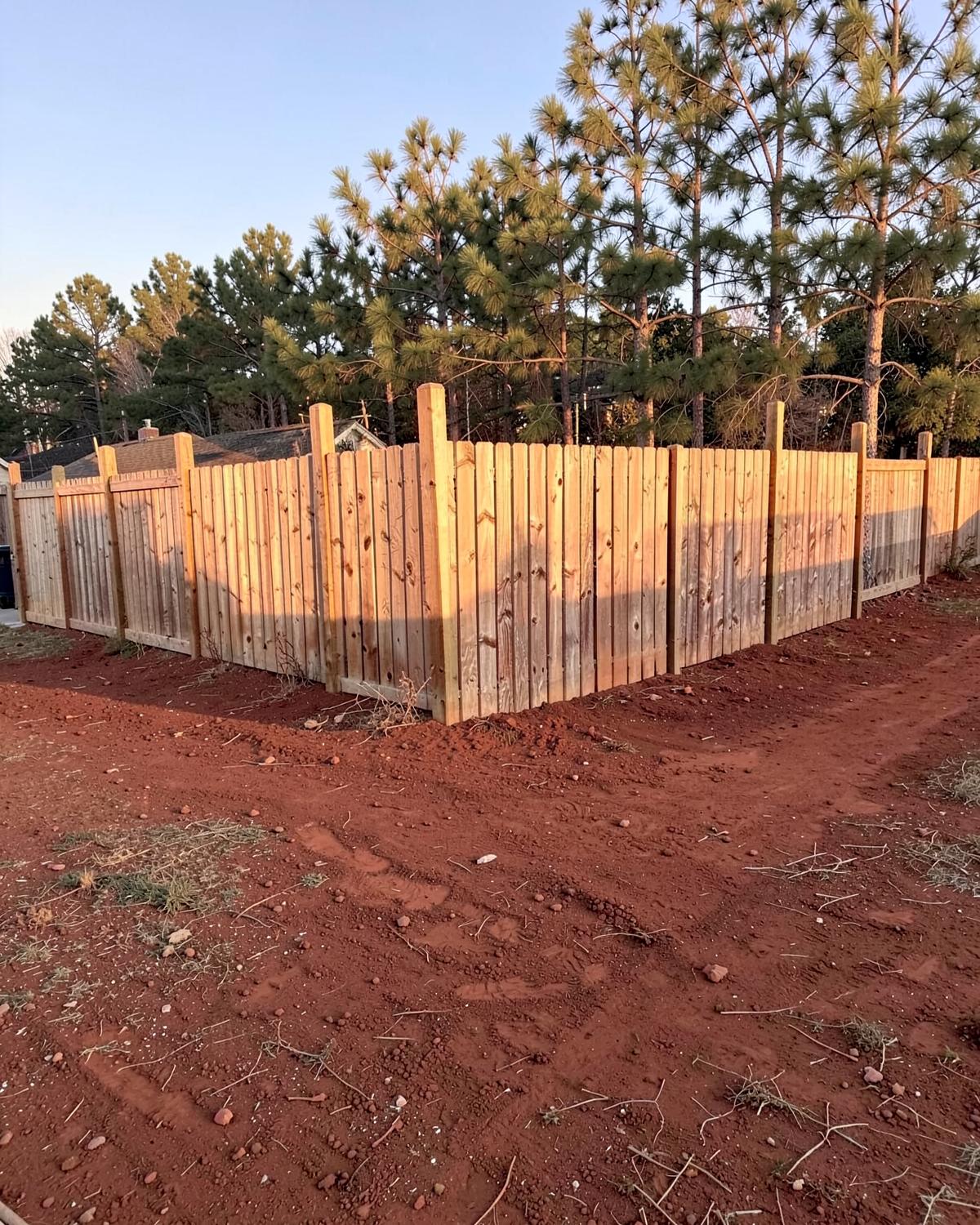 Newly built wooden privacy fence running along the edge of a back yard with fresh red clay soil turned along the post line