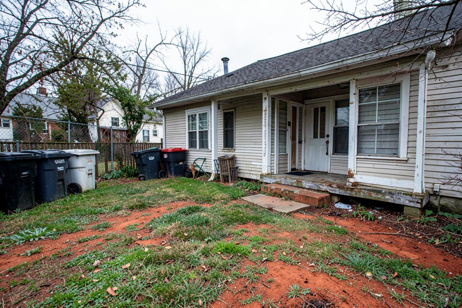 Small back yard of an older southern rental house with no fence, patchy grass, bare red clay and bins beside a worn back stoop