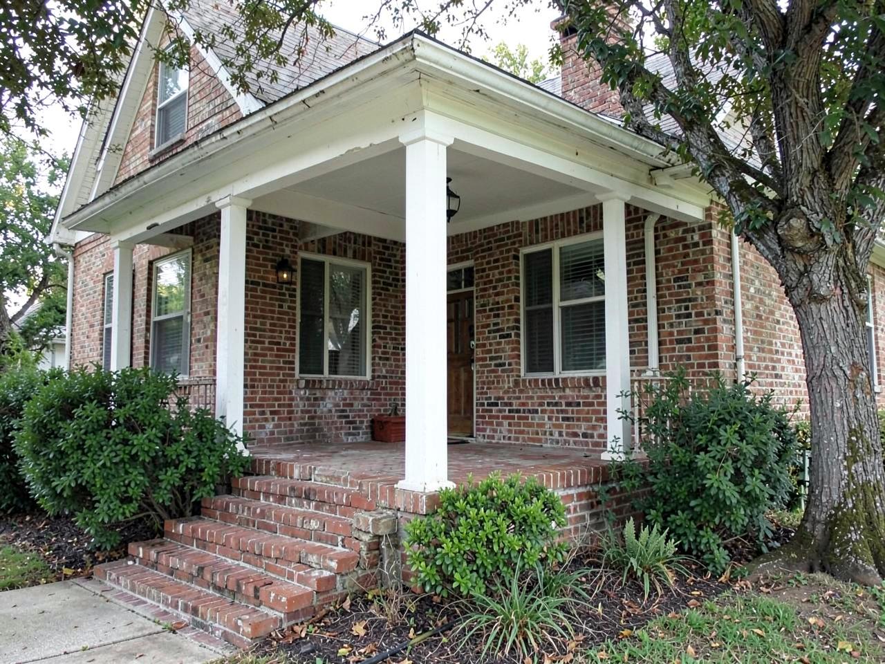 Older brick bungalow with white porch columns and mature trees on a quiet residential street