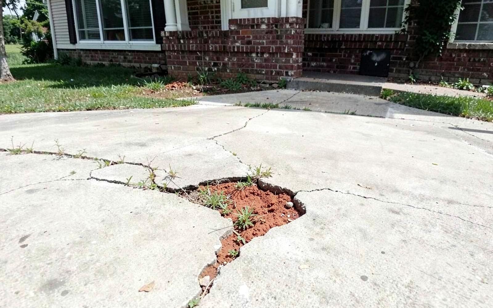 Cracked concrete driveway with a settled seam and red clay soil showing through the gap in front of a brick house