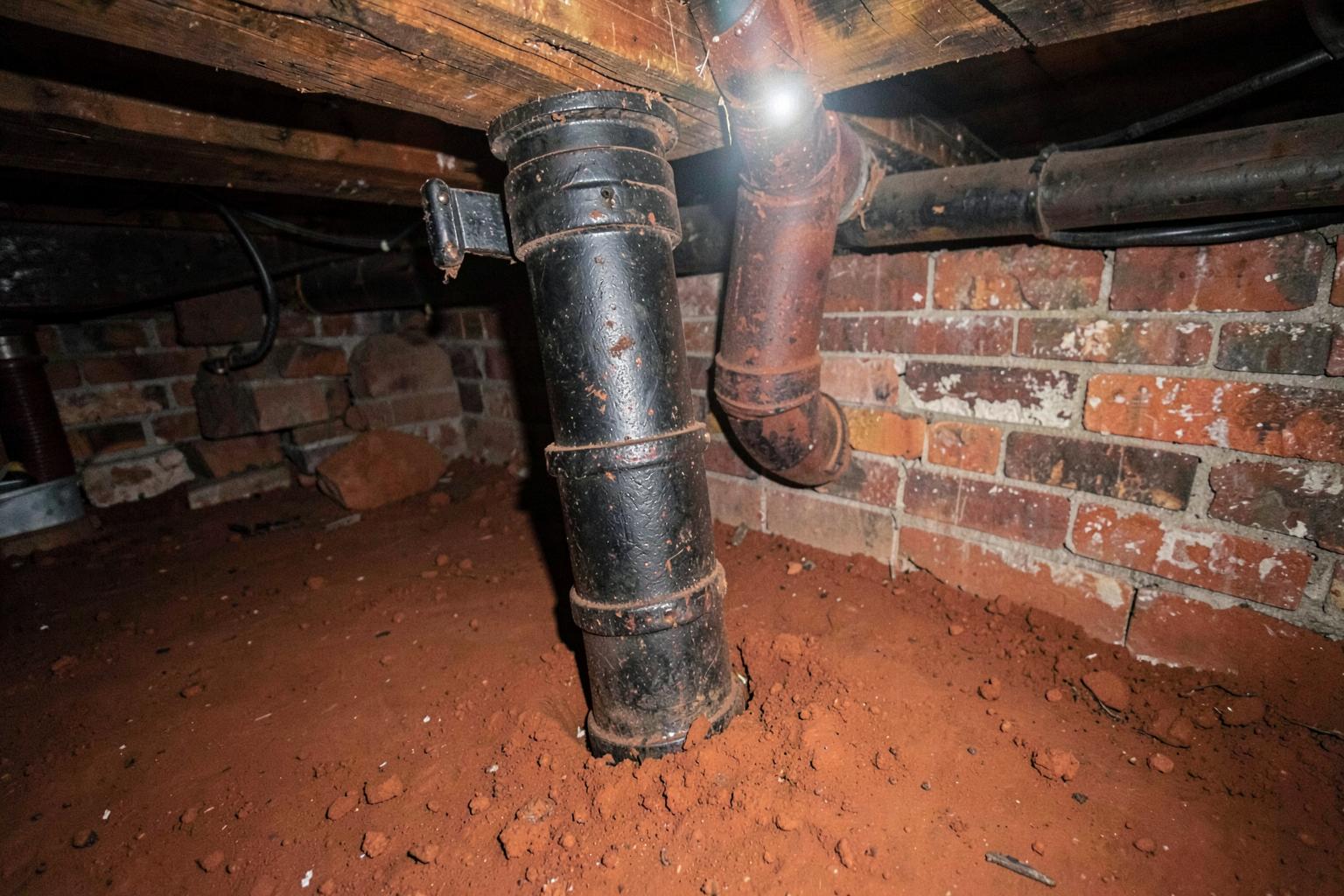 Dim crawlspace under an old house showing brick piers, wooden floor joists and an aged cast iron drain pipe on bare soil