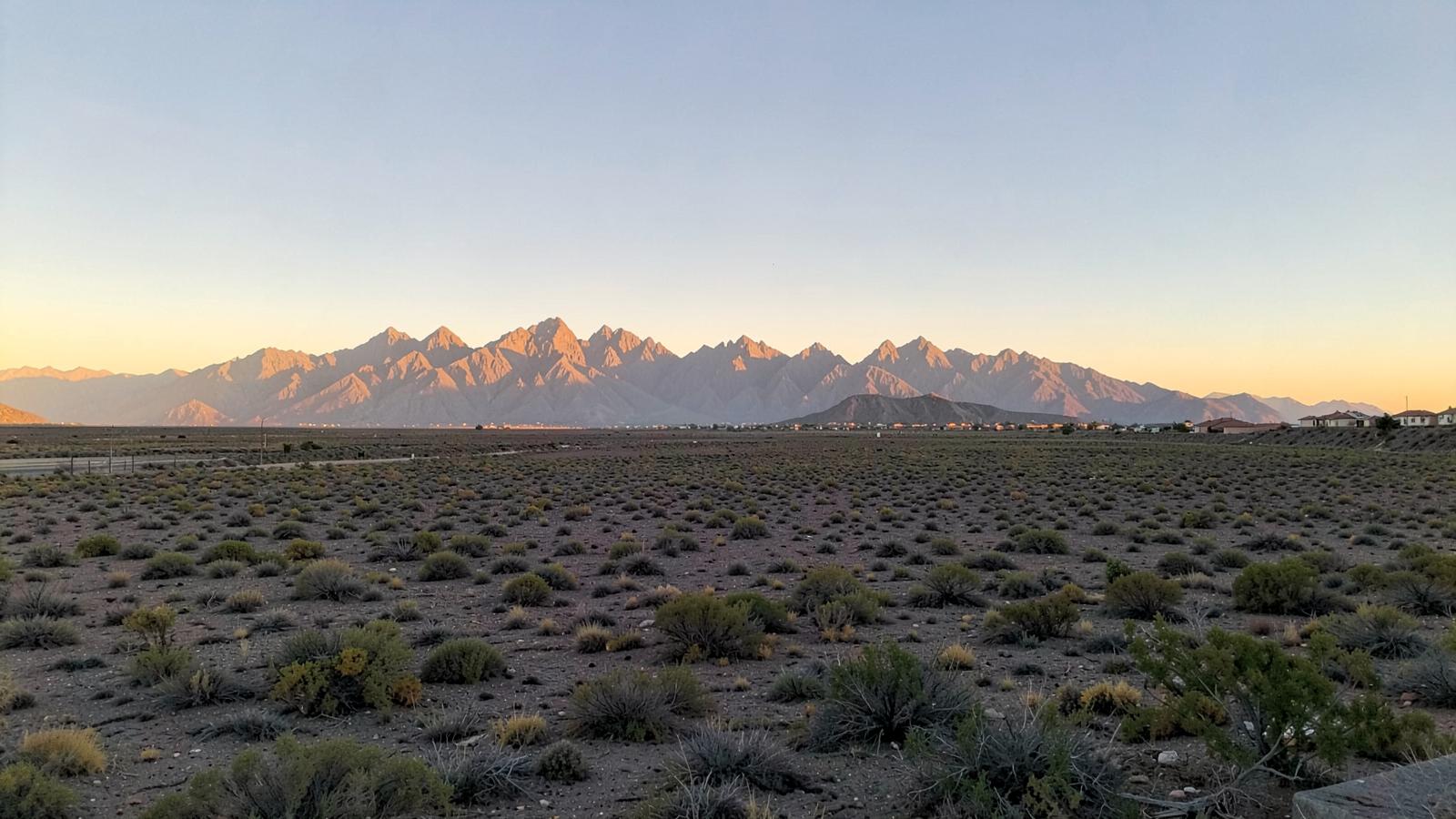 Flat desert basin at sunrise with a jagged mountain range and new rooftops at the far edge