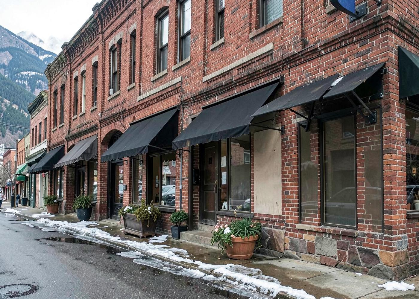 Historic brick storefront block in a mountain town with dark awnings, planters and snow melting along the sidewalk