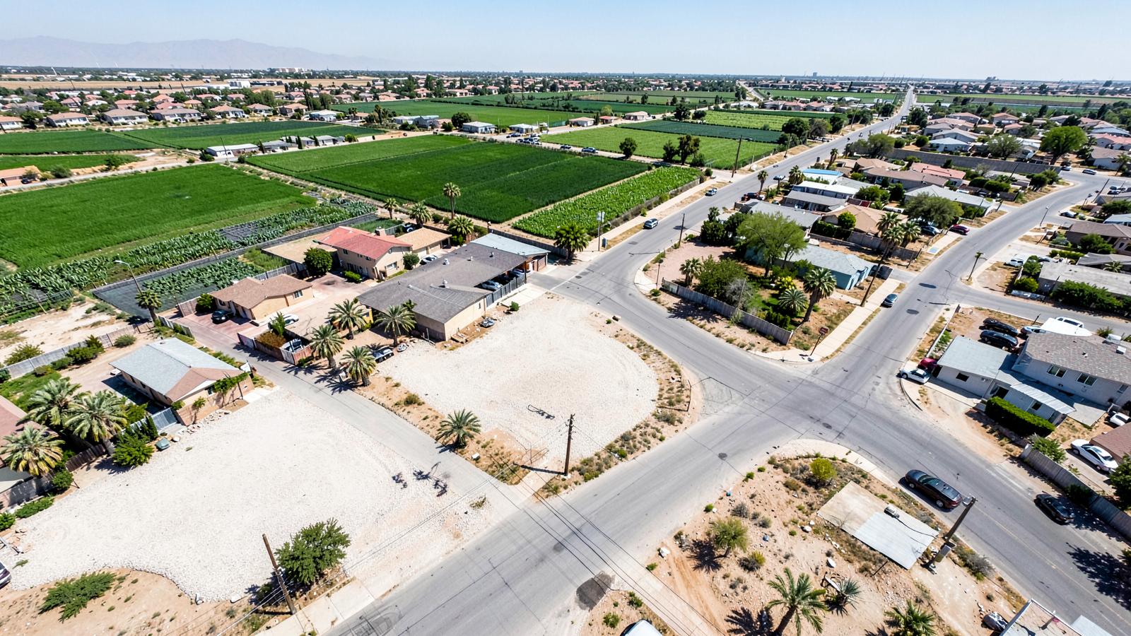 Aerial view of a flat low desert neighbourhood of tile-roof homes bordering irrigated green farm fields under harsh light
