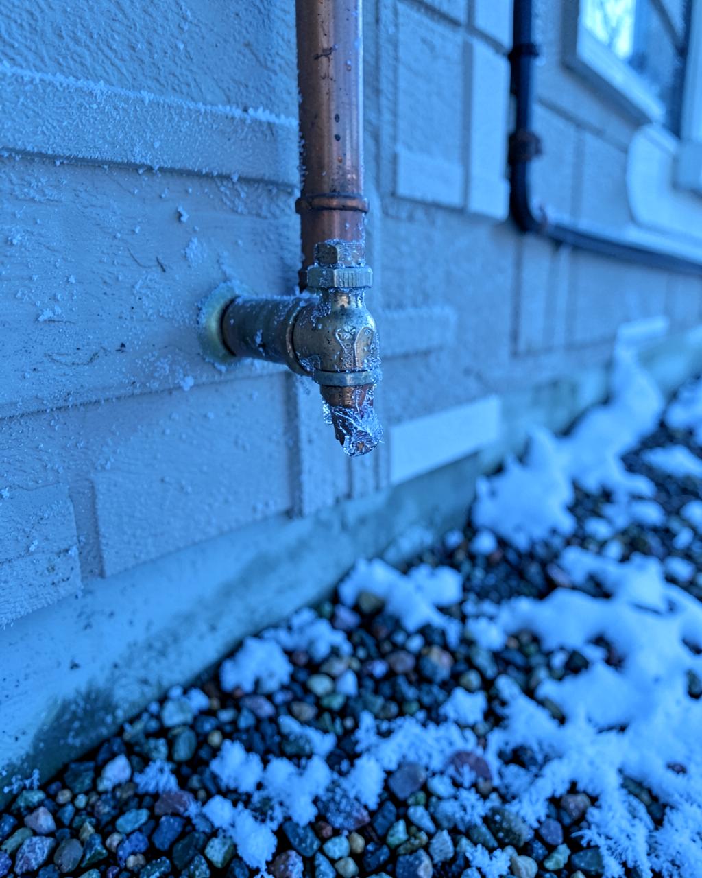 Frost covered outdoor faucet and copper pipe on a house wall above snow dusted gravel in cold morning light