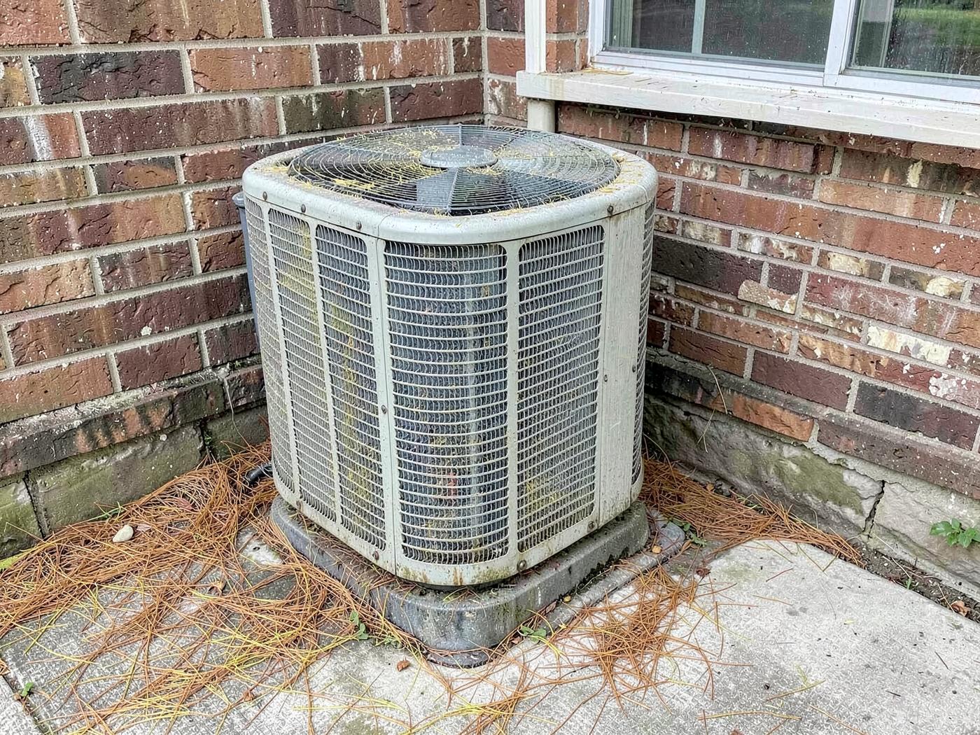 Aged outdoor air conditioning condenser beside a brick house wall with yellow pine pollen dust on the fins and pine needles on the concrete pad