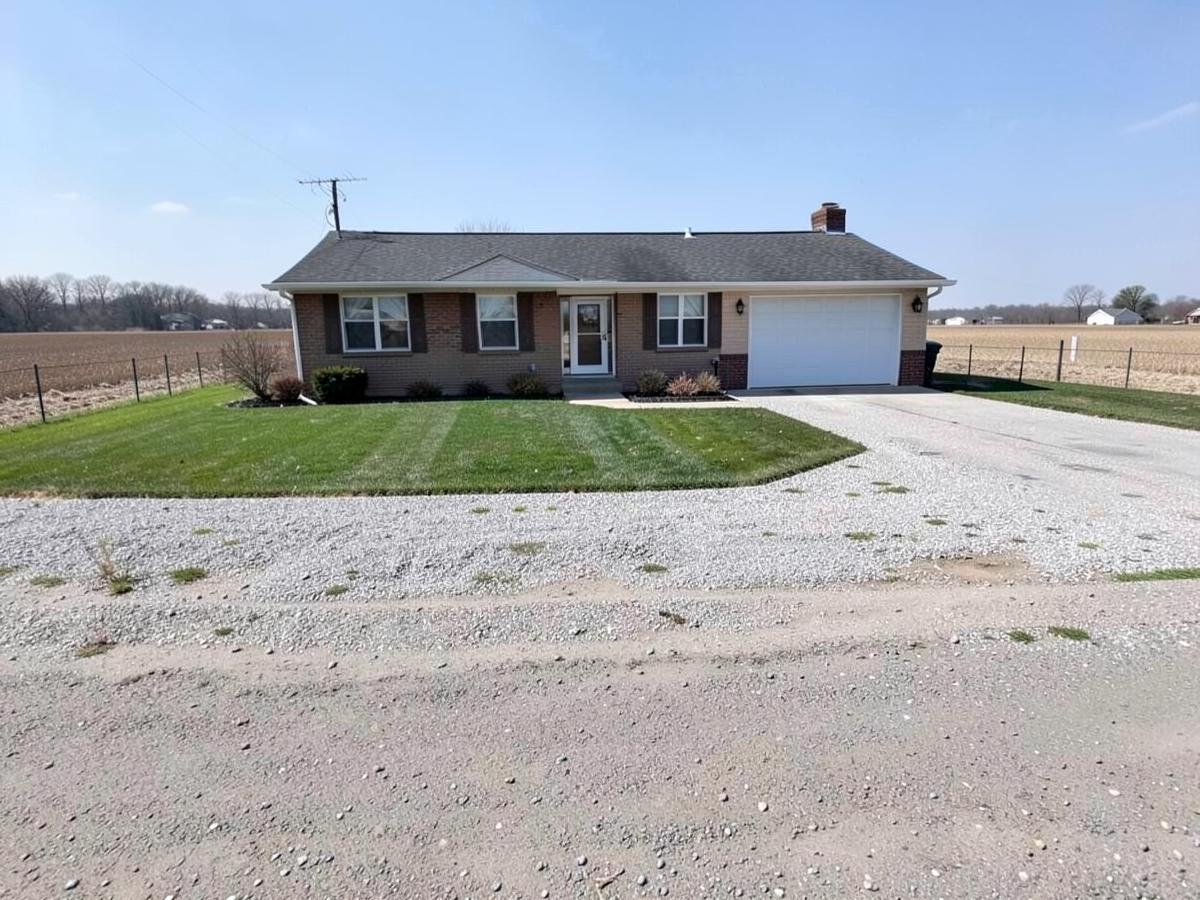 Modest single-story ranch home with a gravel driveway and lawn on a flat street with open farmland at the end of the block