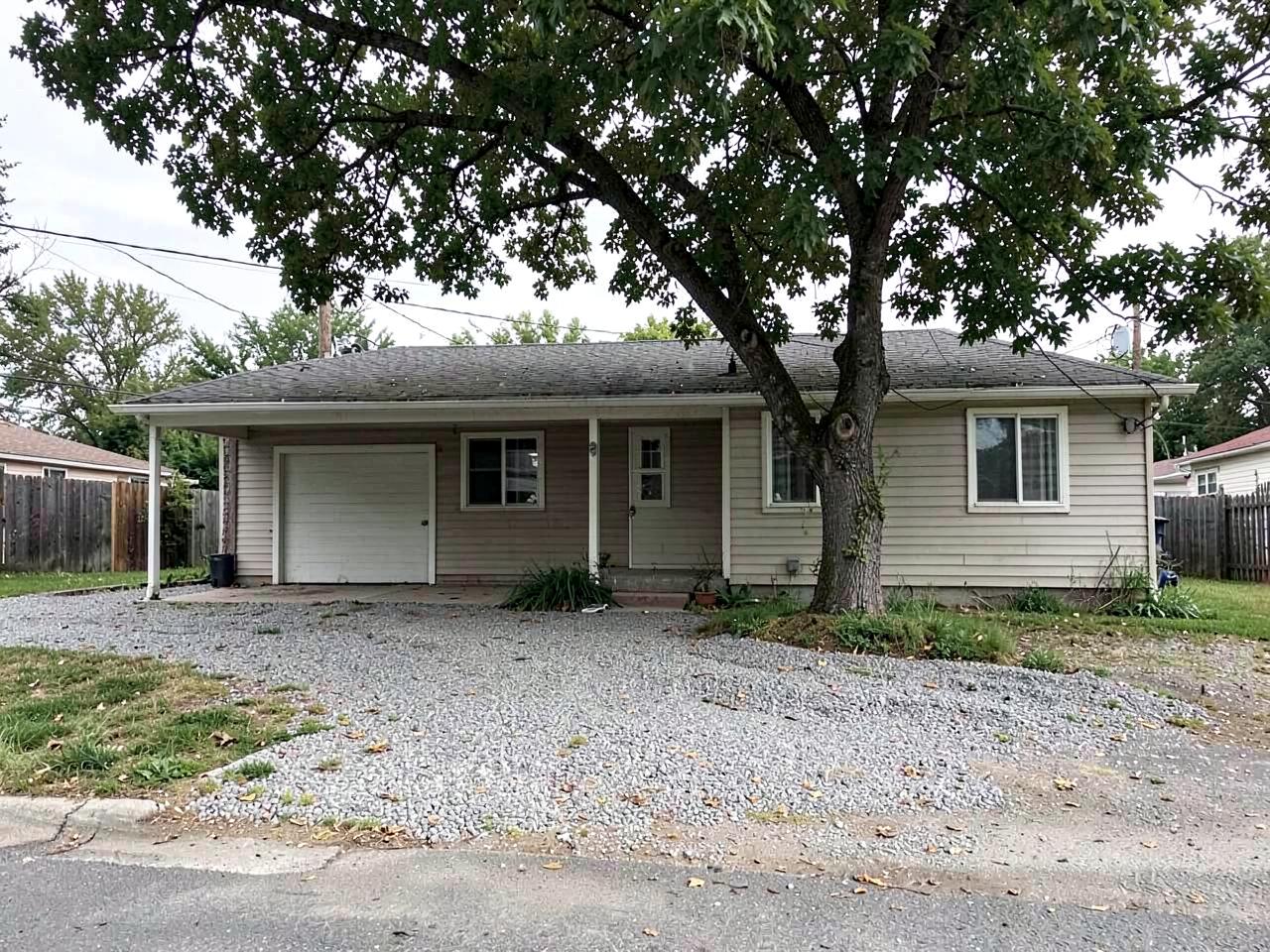 Modest mid-century ranch house with a carport, plain siding and a gravel driveway under a large shade tree