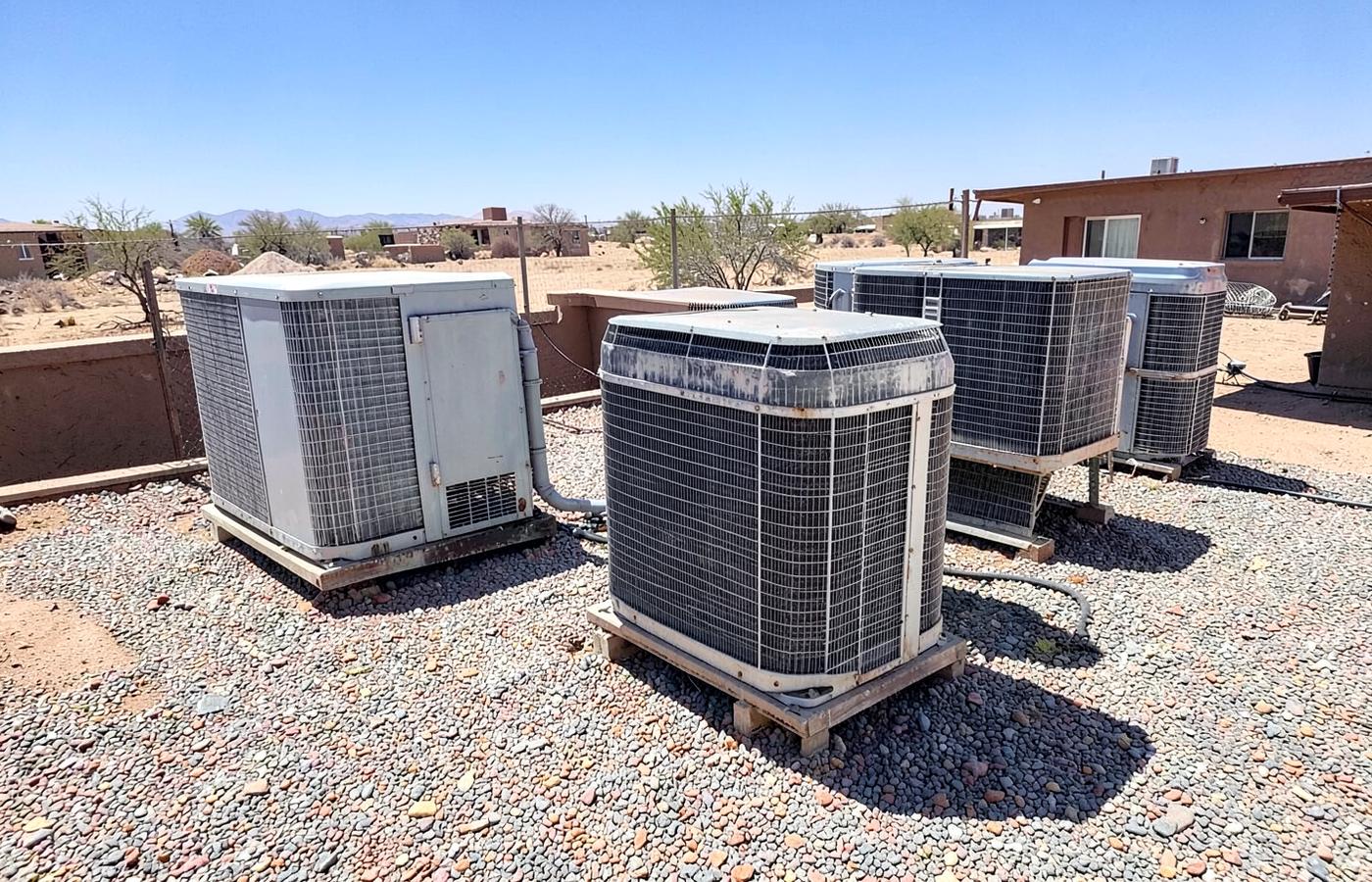 Aging air conditioning condenser units on gravel beside a modest desert home in harsh sunlight