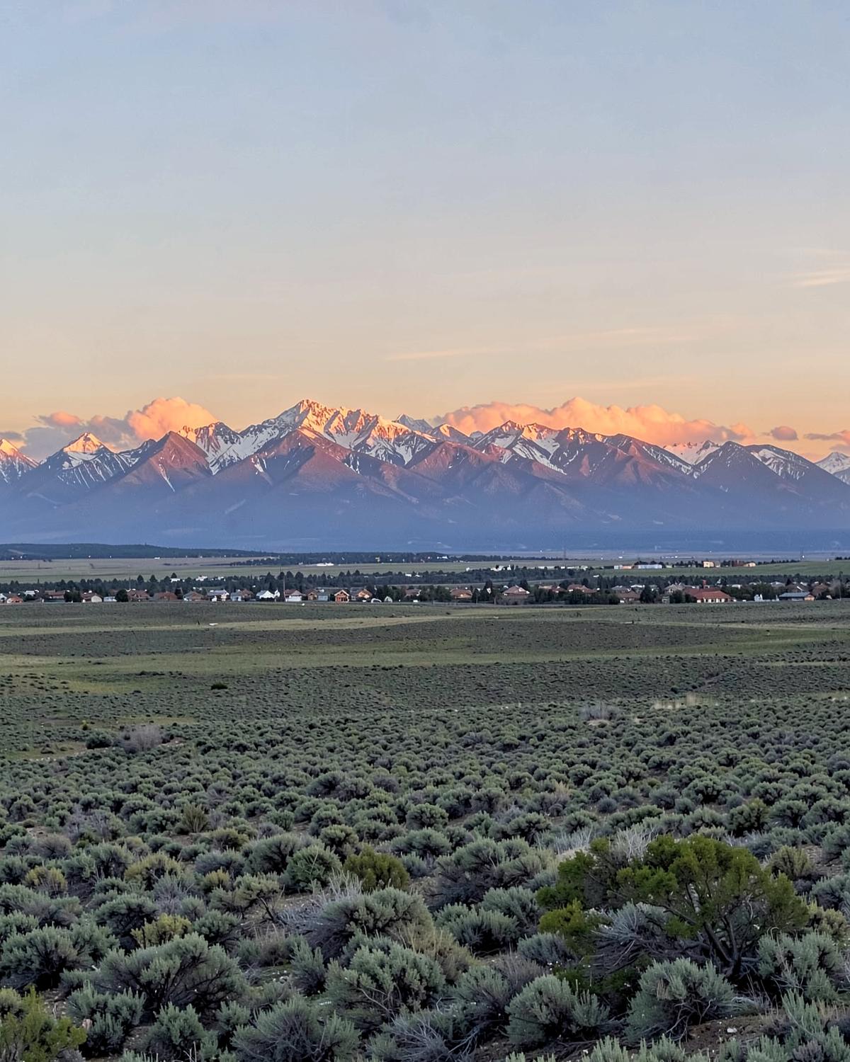 Snow-capped Ruby Mountains rising above a wide sagebrush basin with scattered rooftops far below at golden hour