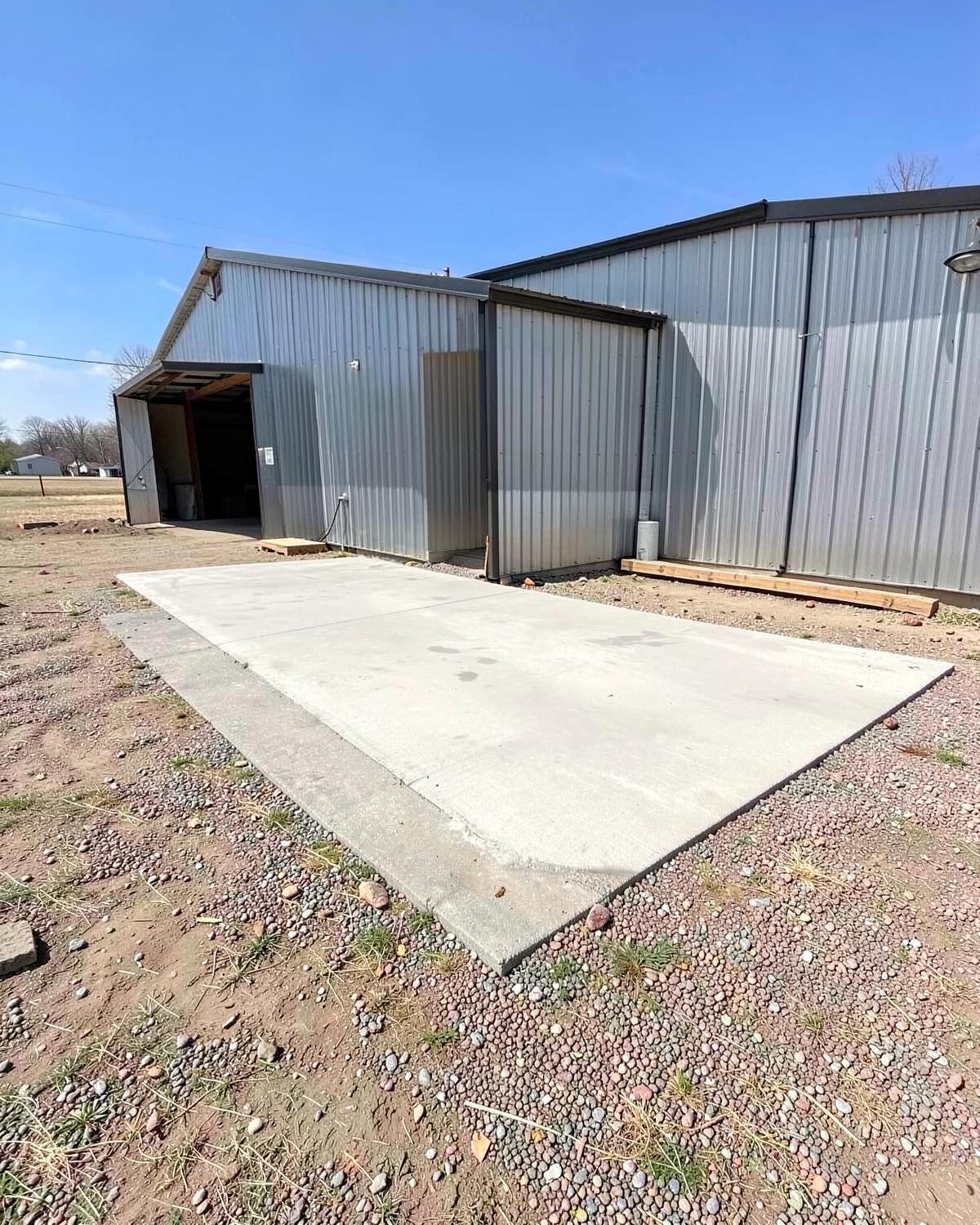 Freshly poured concrete apron in front of a plain metal farm shop building on a gravel lot