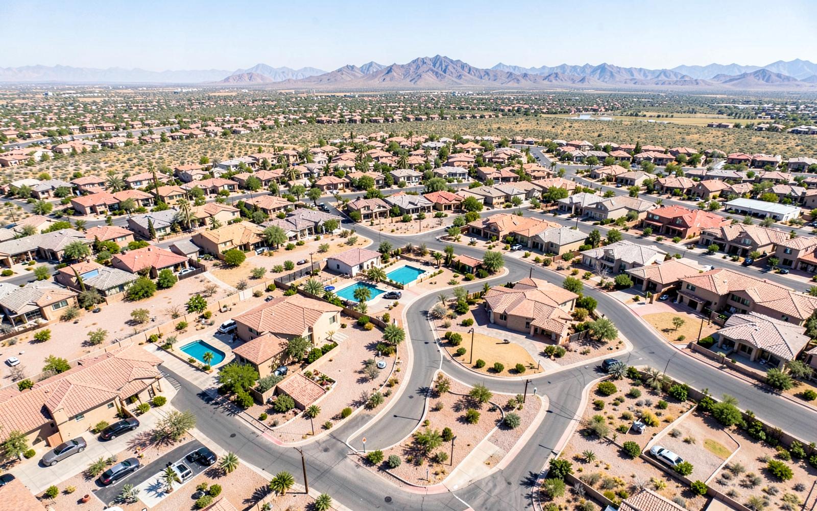 Aerial view of a vast desert suburb with curving streets, tile roofs, backyard pools and gravel yards reaching distant mountains