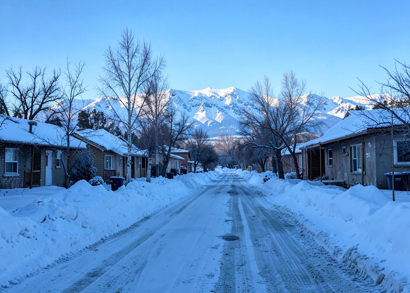 Deep winter residential street in a small high-desert town with plowed snowbanks, frosted roofs and snow-covered mountains beyond