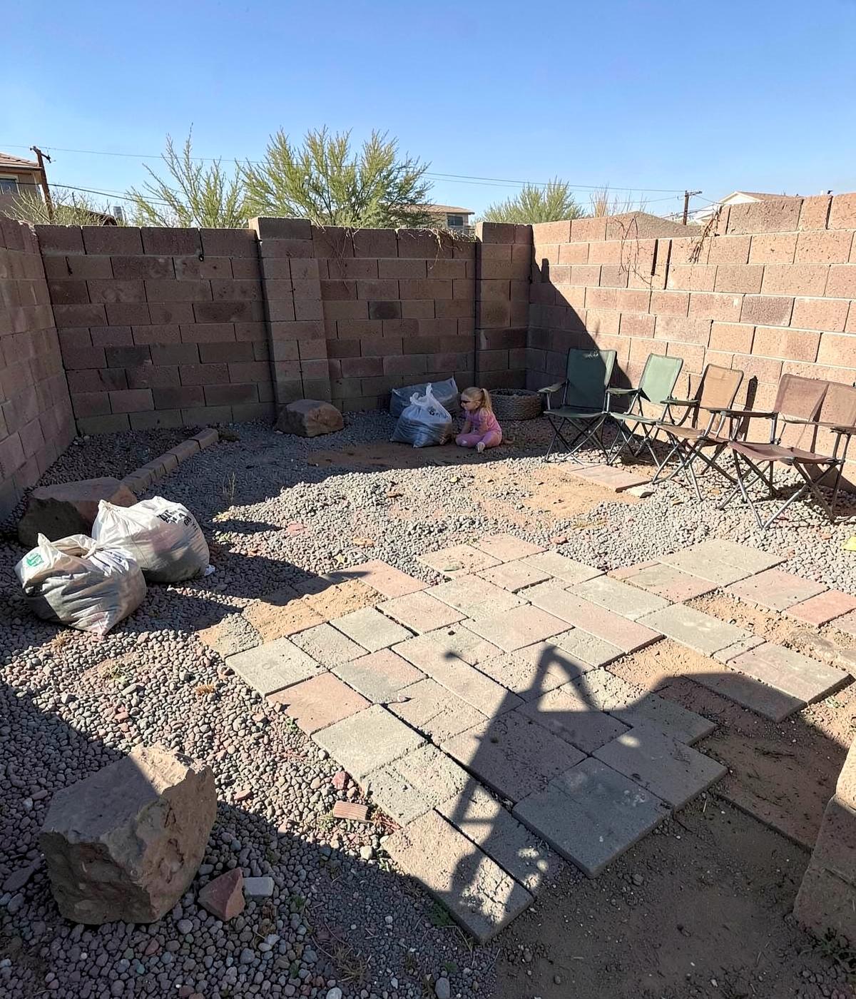 Small desert backyard mid-project with partly laid pavers, gravel, bags of material, and a block wall