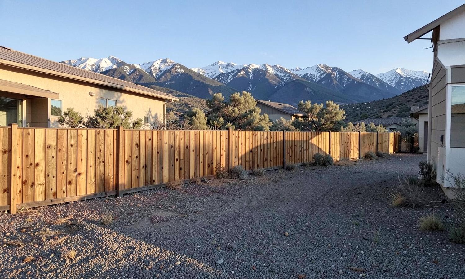 New cedar privacy fence along a gravel side yard of a high-desert home with snow-capped mountains rising behind