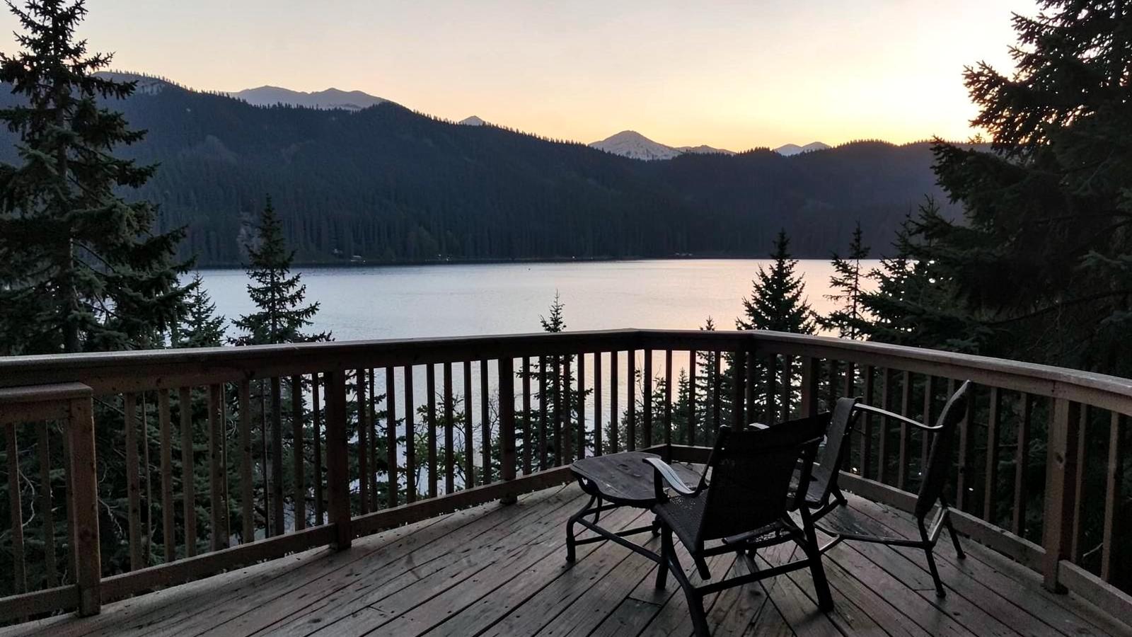 Wooden deck with railing overlooking a forested mountain lake at sunset with outdoor chairs