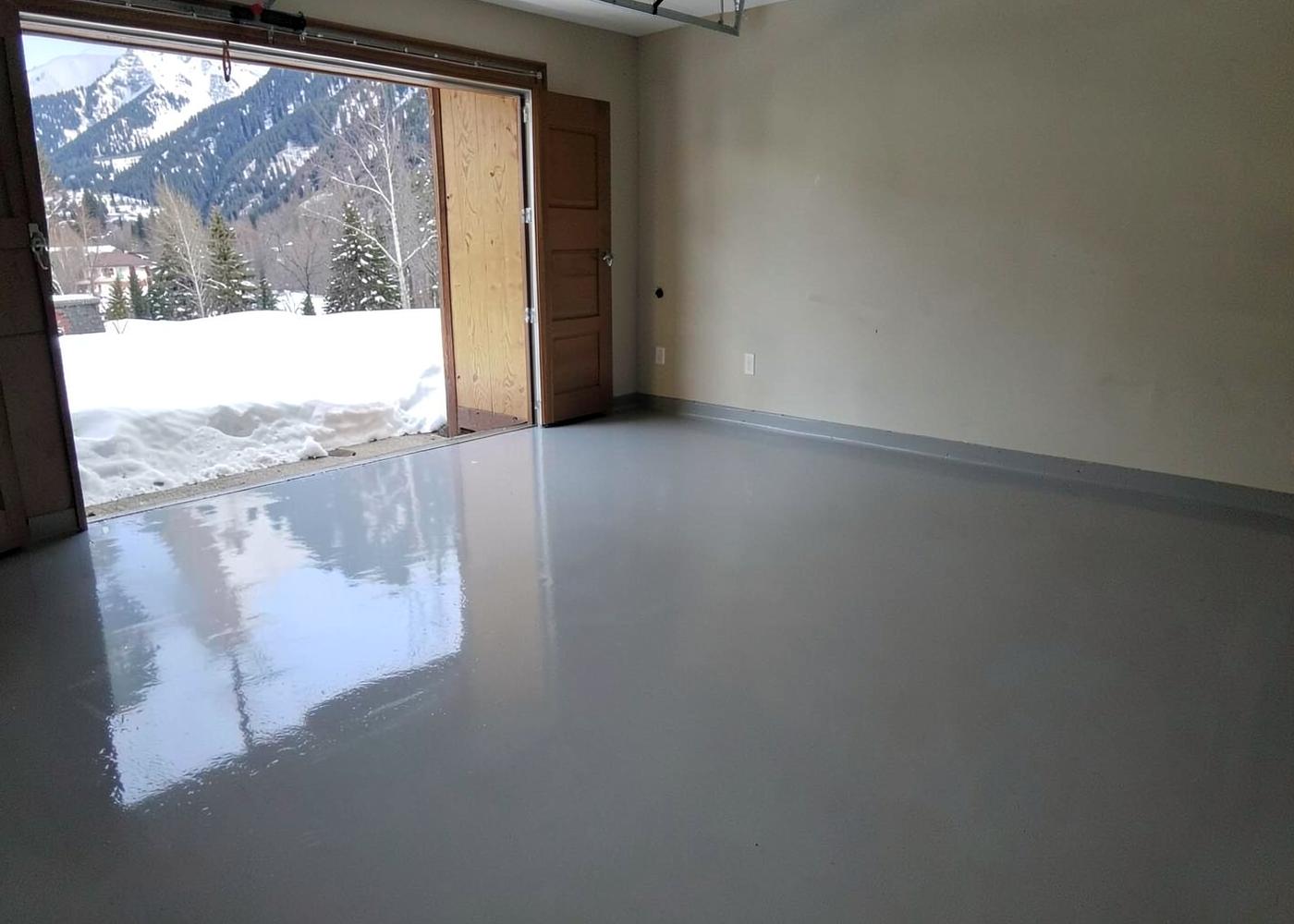 Glossy grey epoxy-coated garage floor in a mountain home with snow and pines visible through the open door
