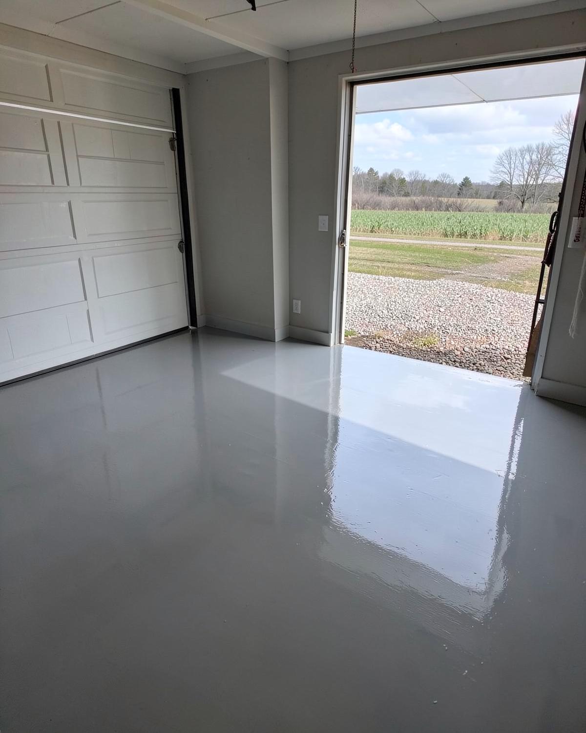 Glossy grey epoxy coated garage floor in a clean garage with the door open to a gravel drive and open country