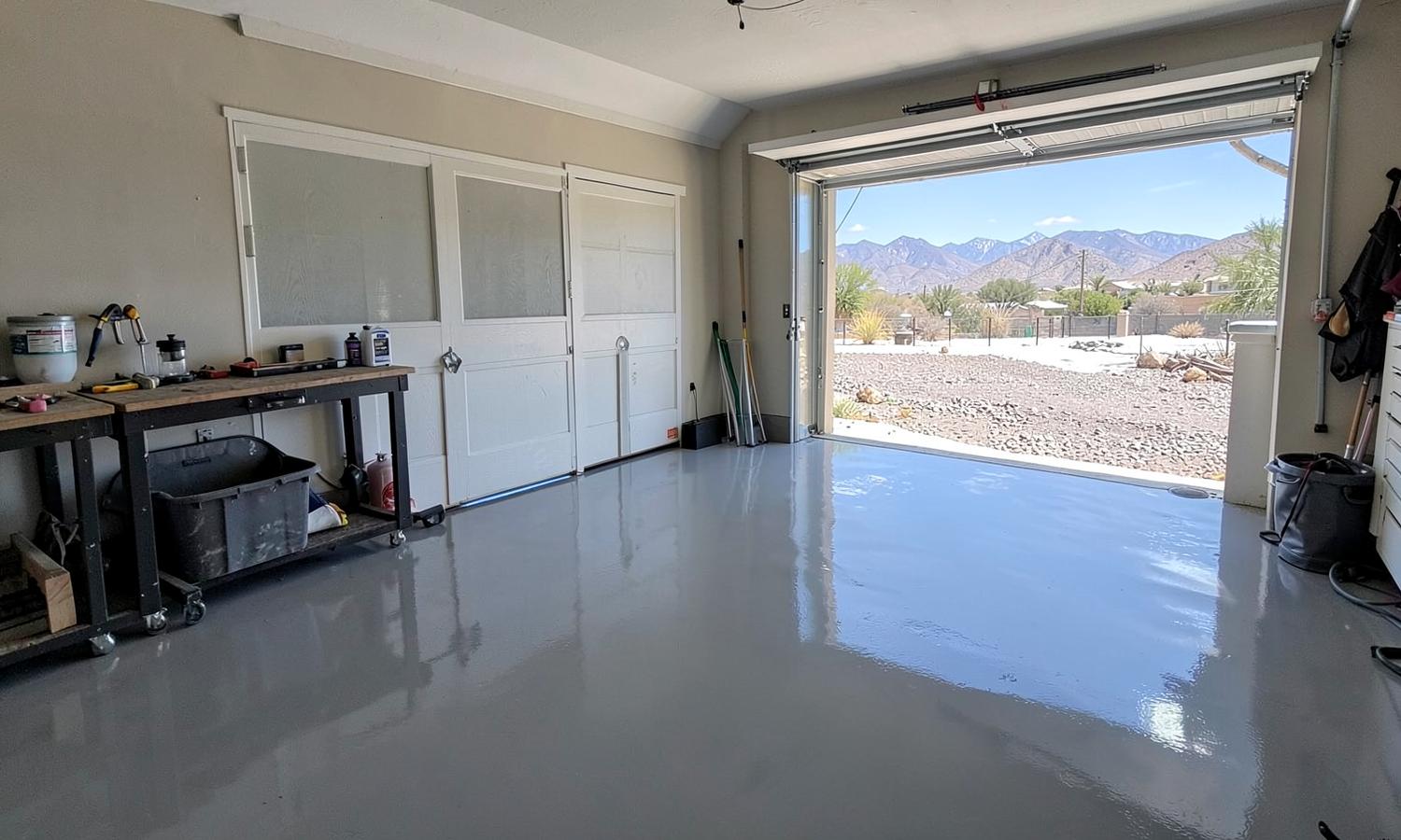 Freshly coated glossy grey epoxy garage floor with a work bench and an open garage door showing a gravel yard and mountains
