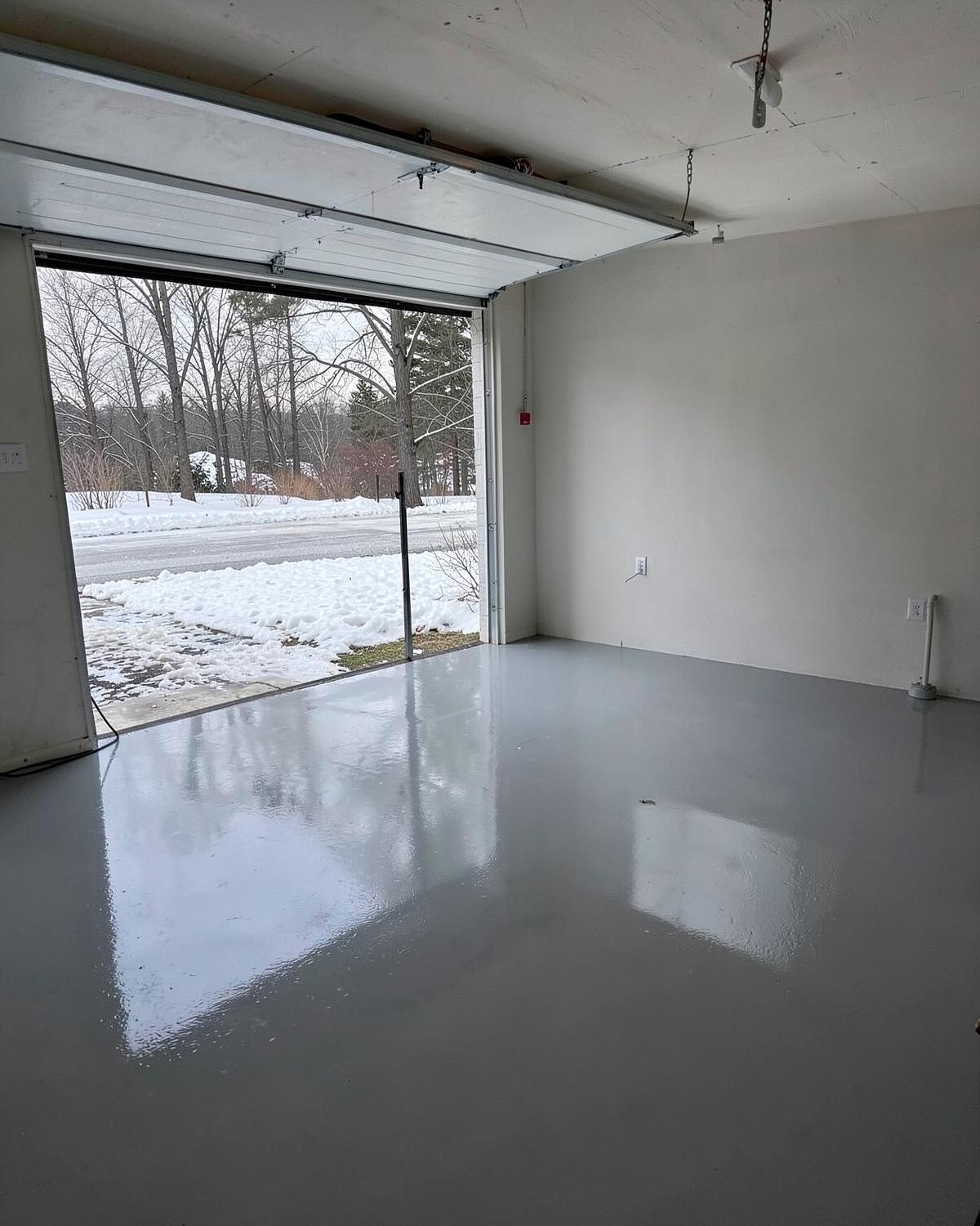 Glossy grey epoxy coated garage floor with the door open onto a snowy driveway and bare trees