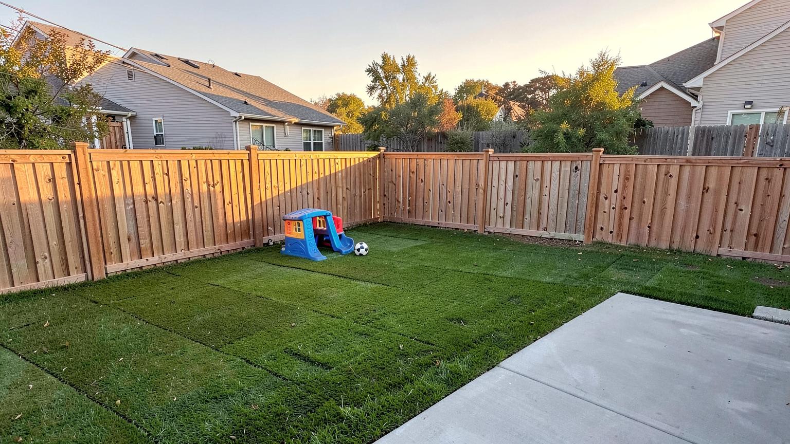 Finished backyard of a new suburban home with fresh sod, patio slab, fence line and a swing set at golden hour