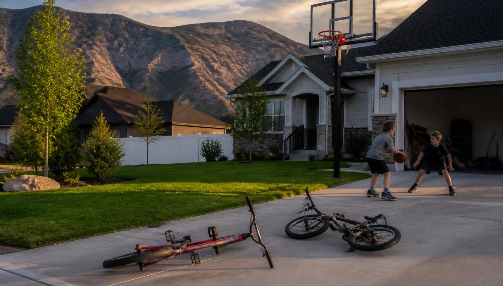 Children playing basketball on the concrete driveway of a newer suburban home with bikes on the ground and mountains behind