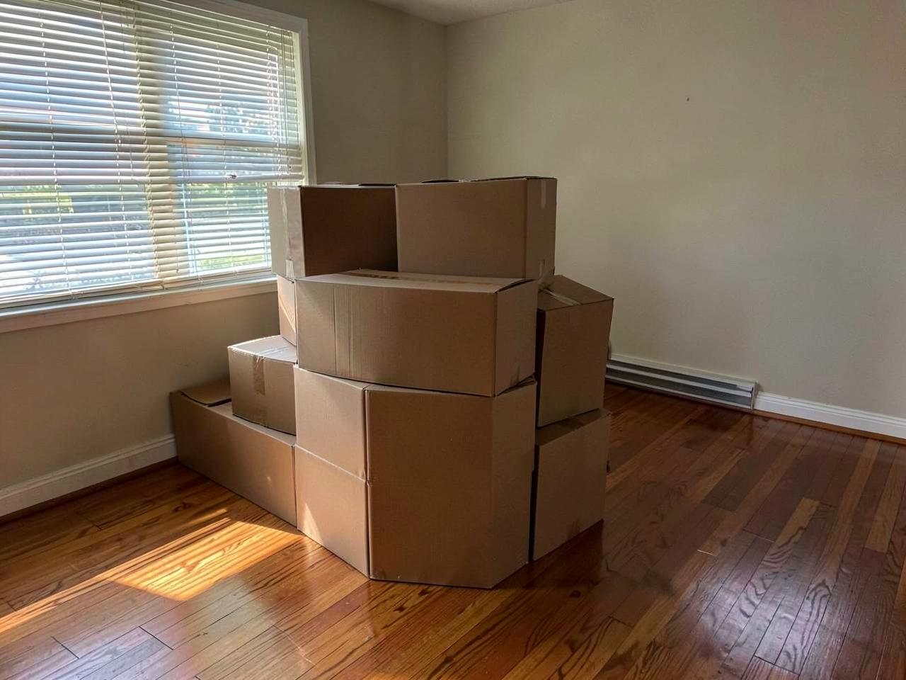 Plain cardboard moving boxes stacked in an empty living room with hardwood floors and afternoon light through blinds