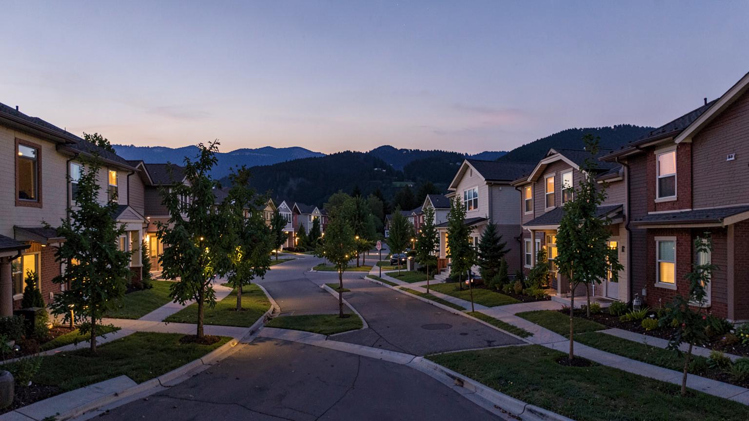 New suburban neighborhood street at dusk with warm windows, young trees and mountains silhouetted behind