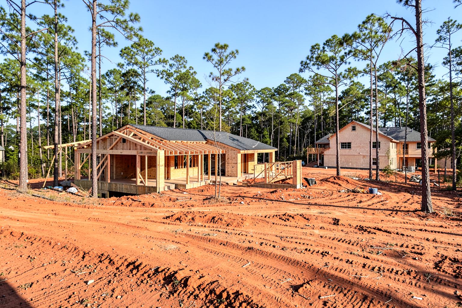 New wood-framed houses under construction on red clay lots surrounded by tall standing pines