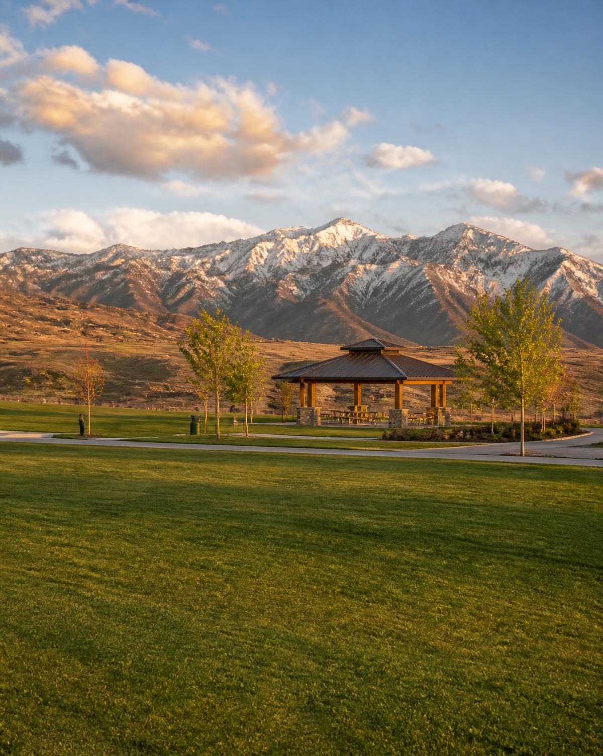 Empty community park with mown grass, a pavilion and young trees below a snow-dusted mountain range at golden hour