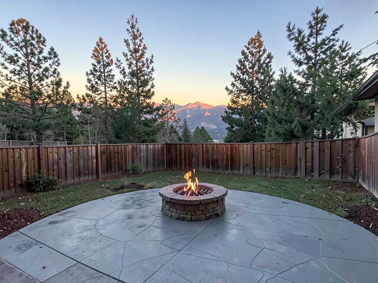 Backyard stamped concrete patio with a lit stone fire pit, tall pines, and a mountain ridge at sunset