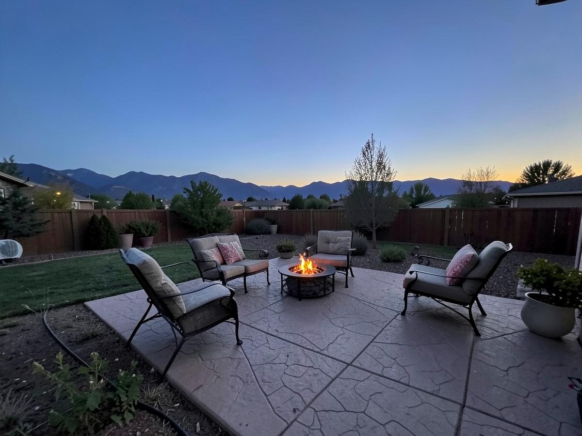 Stamped concrete patio with outdoor chairs and a lit fire pit in a wide backyard at dusk with mountains on the horizon