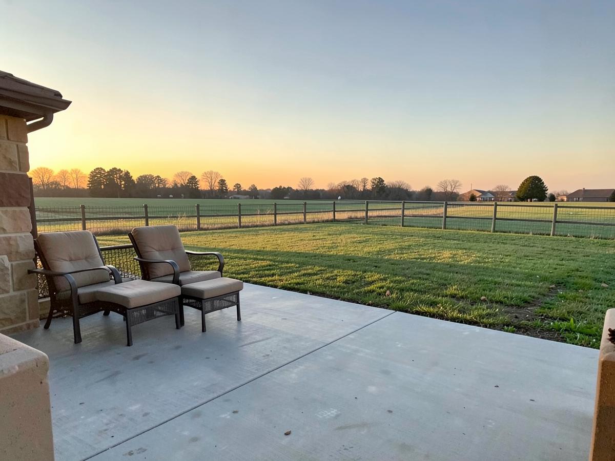 Backyard concrete patio with simple outdoor chairs and a lawn at golden hour with open farm country beyond the fence