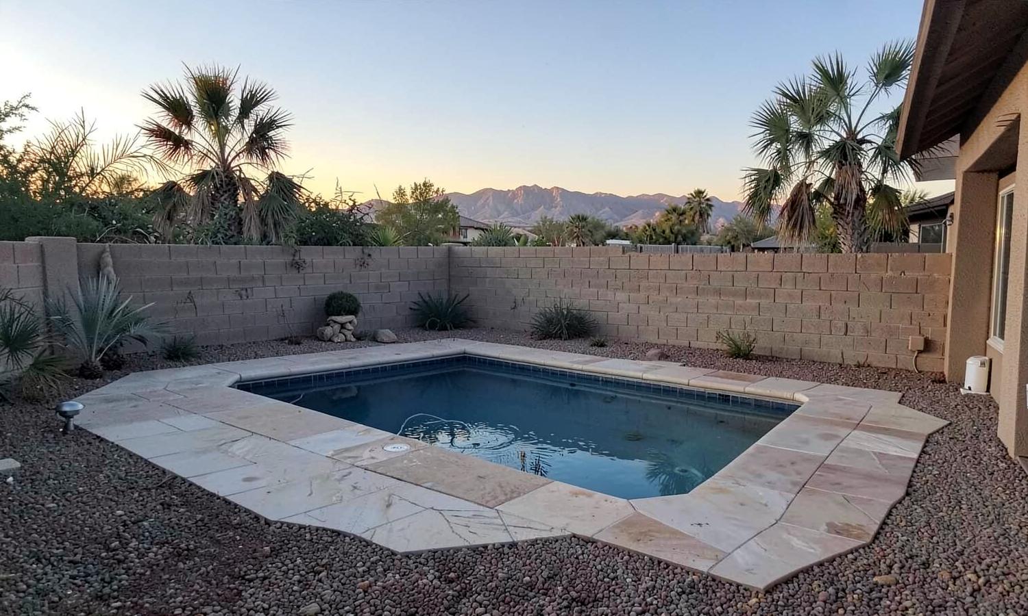 Small backyard swimming pool with a travertine deck, block wall, and palms with desert mountains beyond at dusk