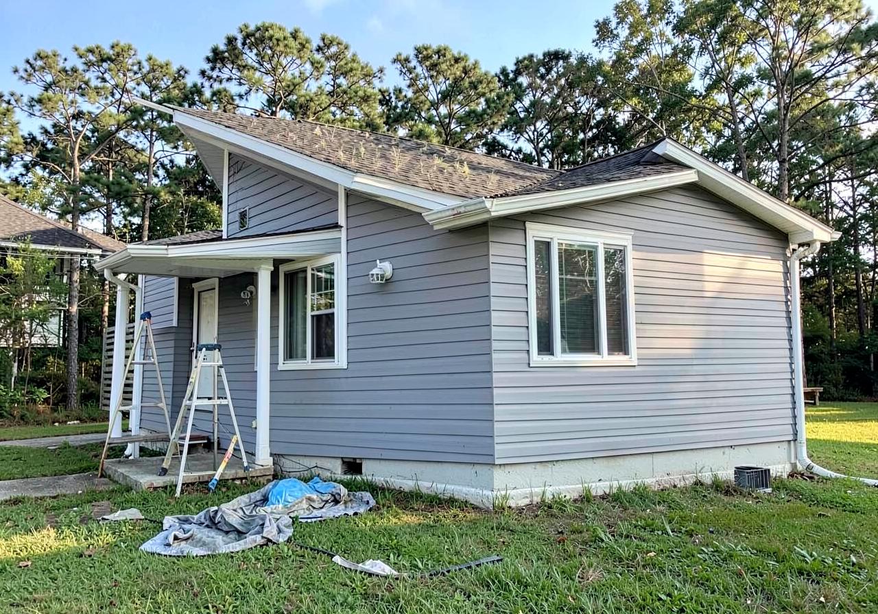 Suburban house freshly repainted in soft gray with white trim, ladder and drop cloth on the lawn, pines behind