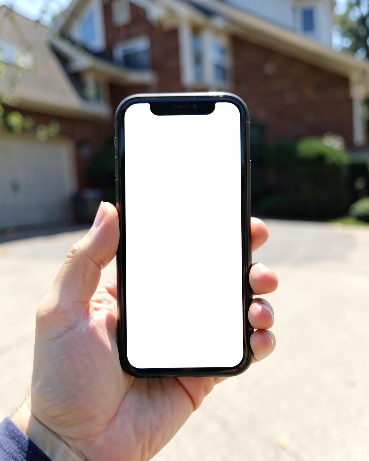 Hand holding a smartphone with a blank white screen in front of a residential driveway in bright daylight