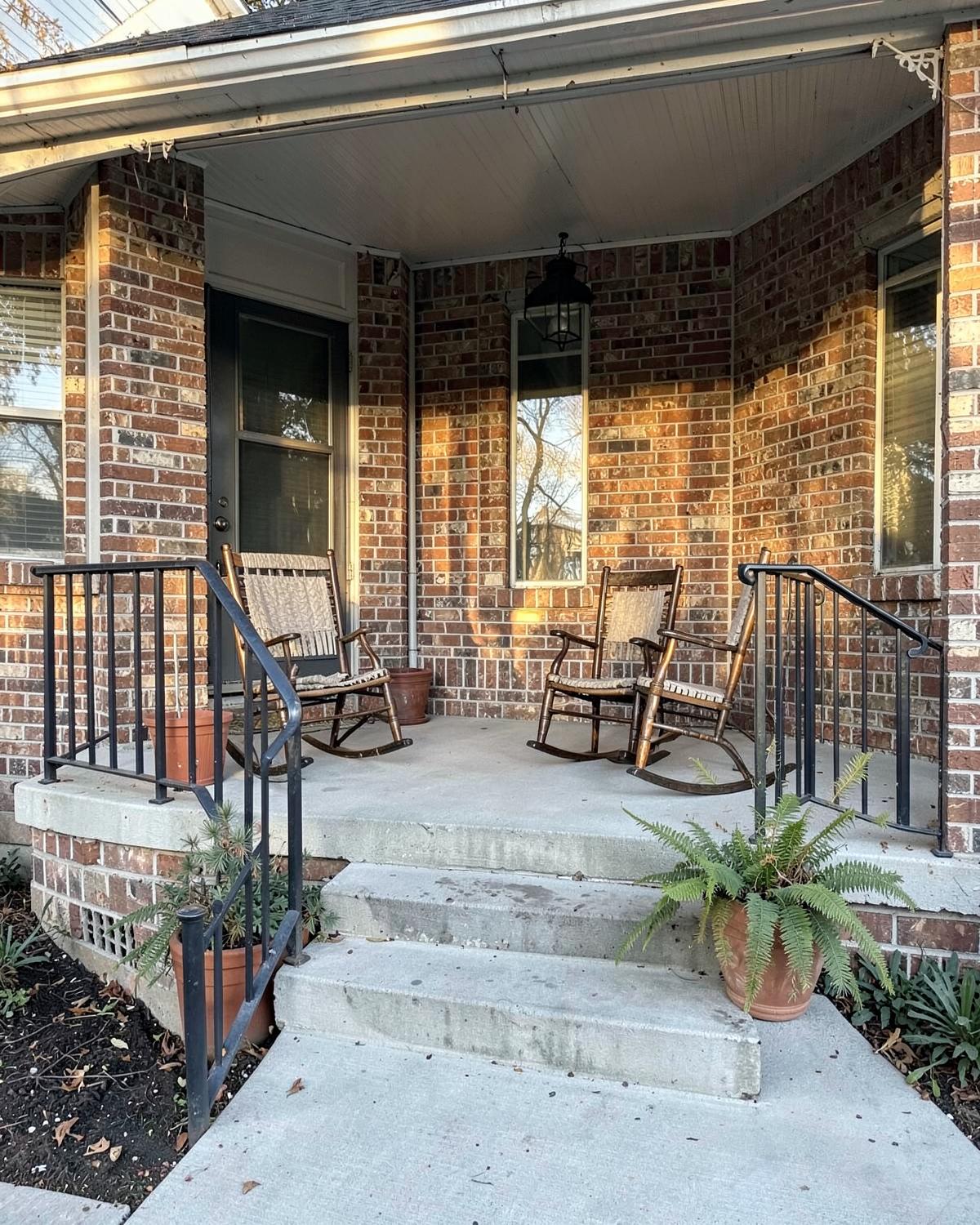 Two rocking chairs on the shaded brick porch of an older Columbus home with mature pines beyond the railing