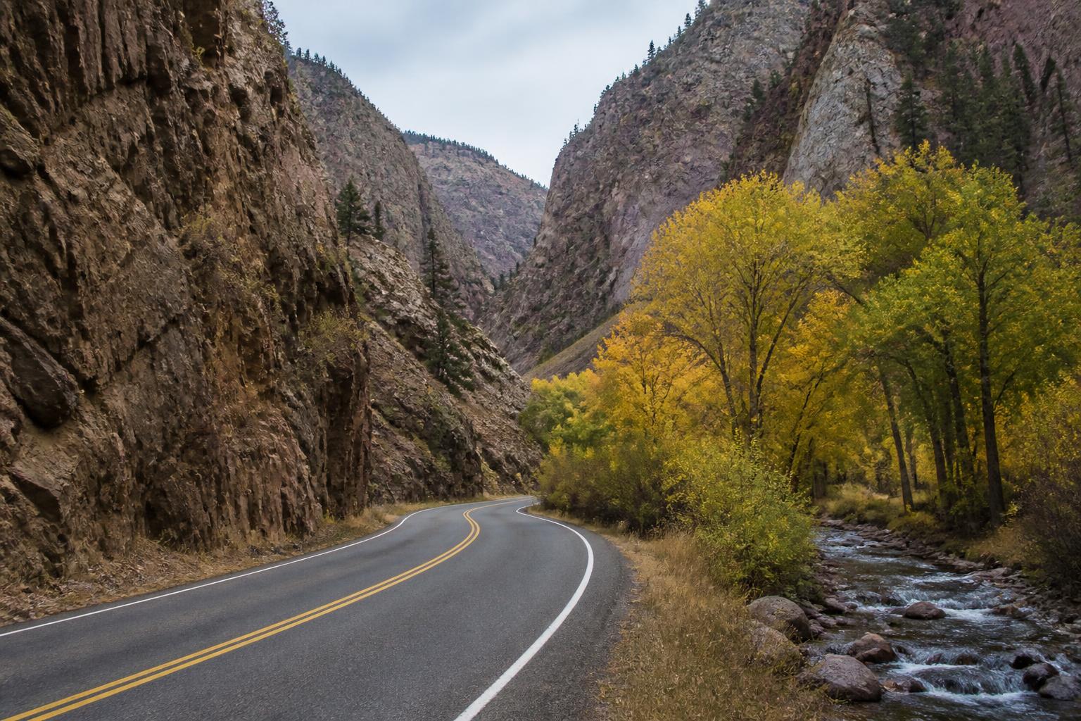 Two-lane canyon road curving between steep rocky walls beside a creek and yellow cottonwoods