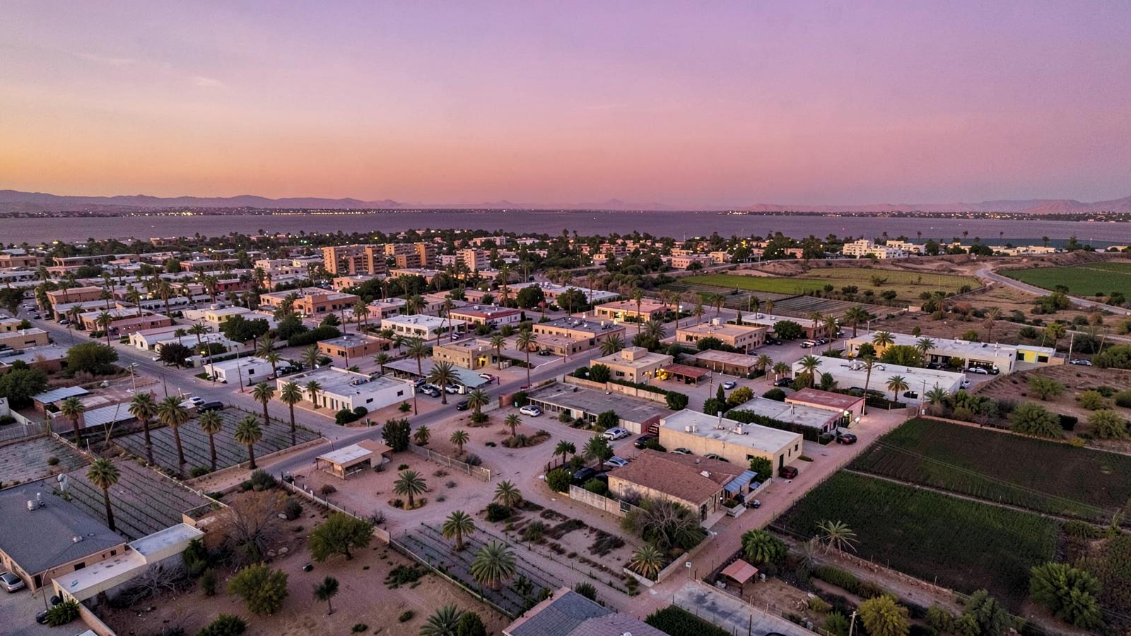 Aerial view at dusk of a low desert city grid of palms and low buildings with irrigated fields at its edge under an amber sky