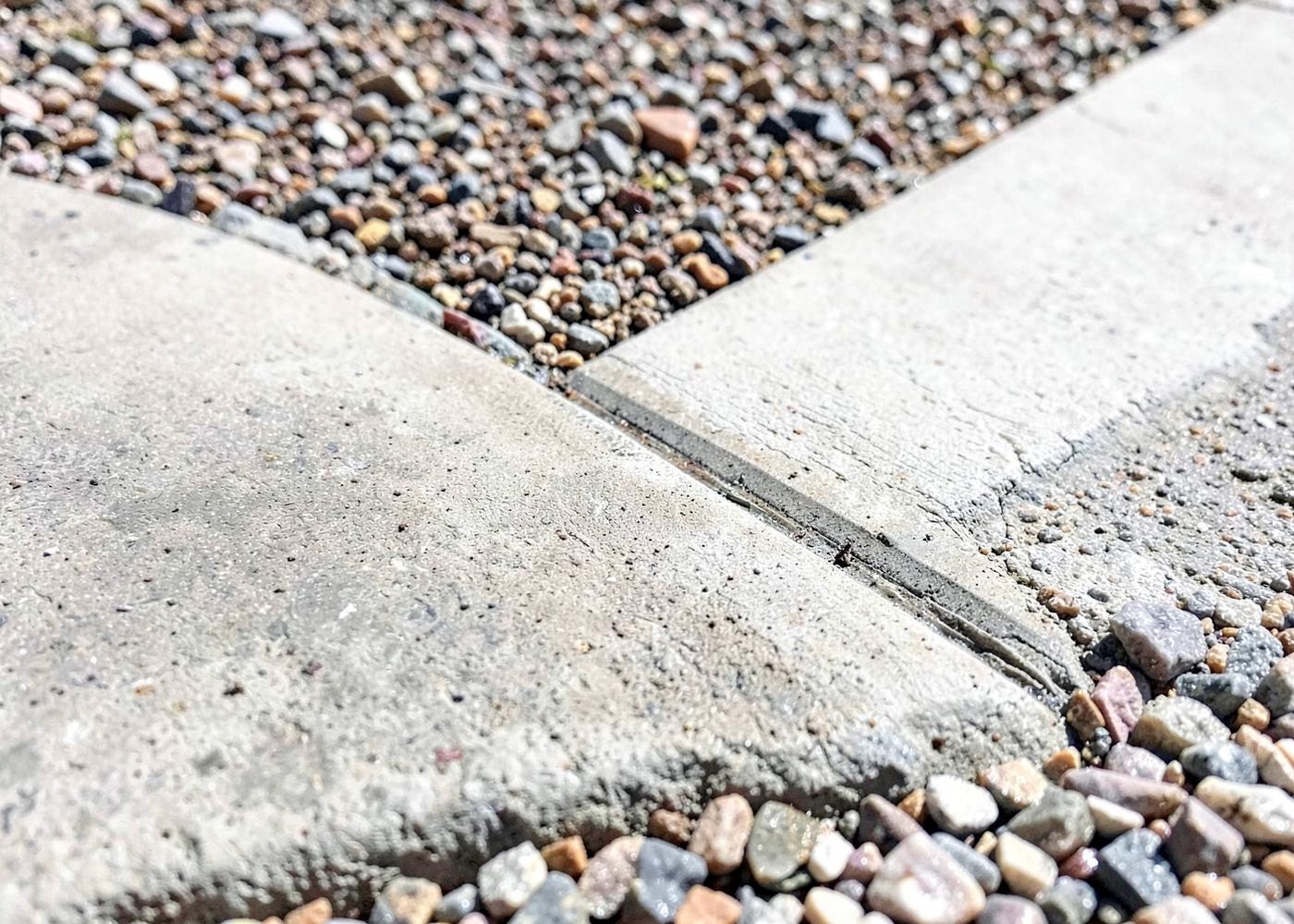 Close detail of a freshly finished concrete driveway edge meeting desert gravel with a crisp control joint in strong sunlight